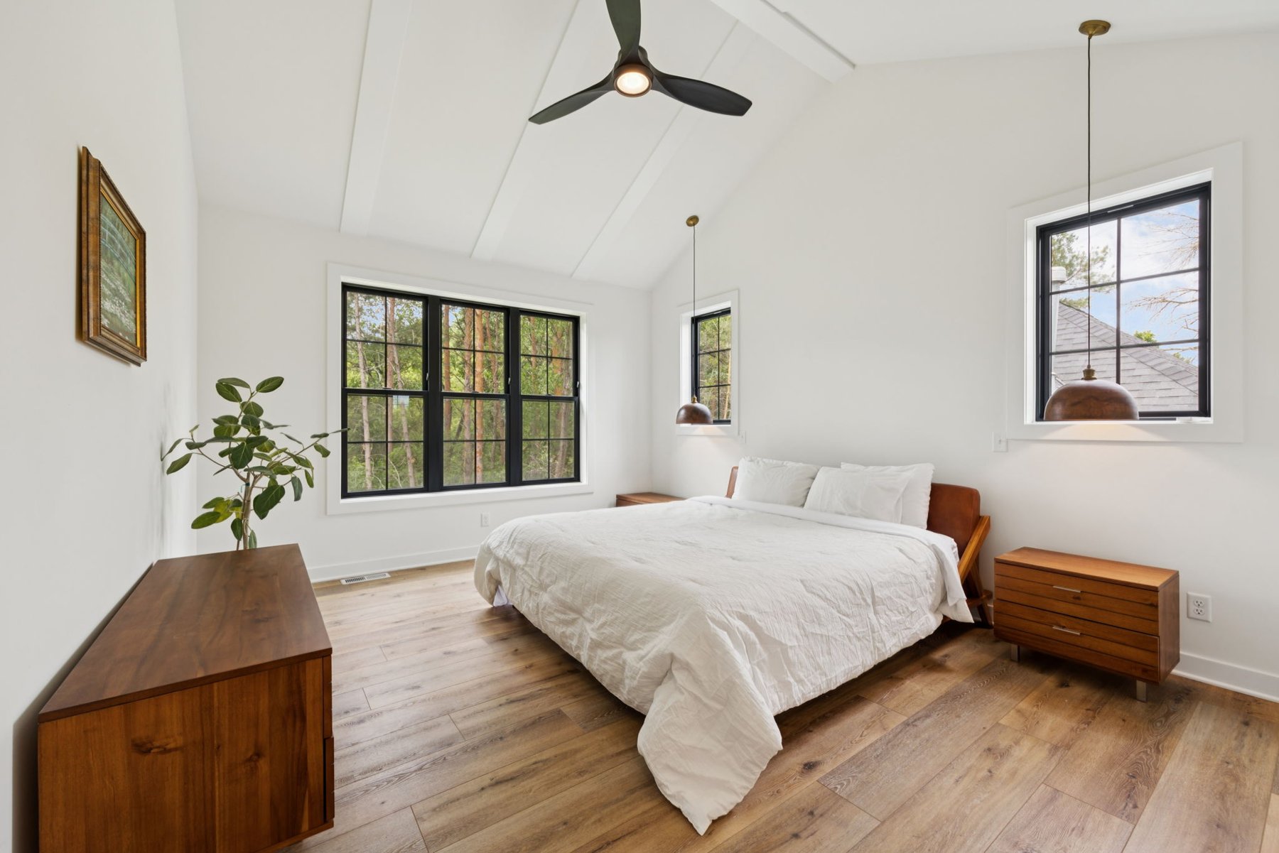 Main-level primary bedroom with a vaulted ceiling, wide-plank oak floors, and black-framed windows looking into the woods